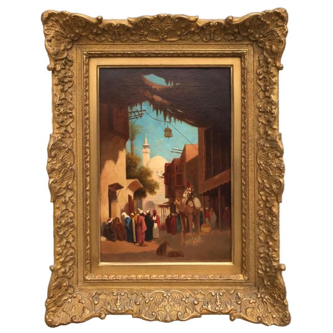 Huile sur panneau de Théodore Frère représentant une scène de rue animée au Caire, avec figures en habits traditionnels, architecture orientale et minaret à l’arrière-plan, dans un cadre en bois doré richement sculpté.